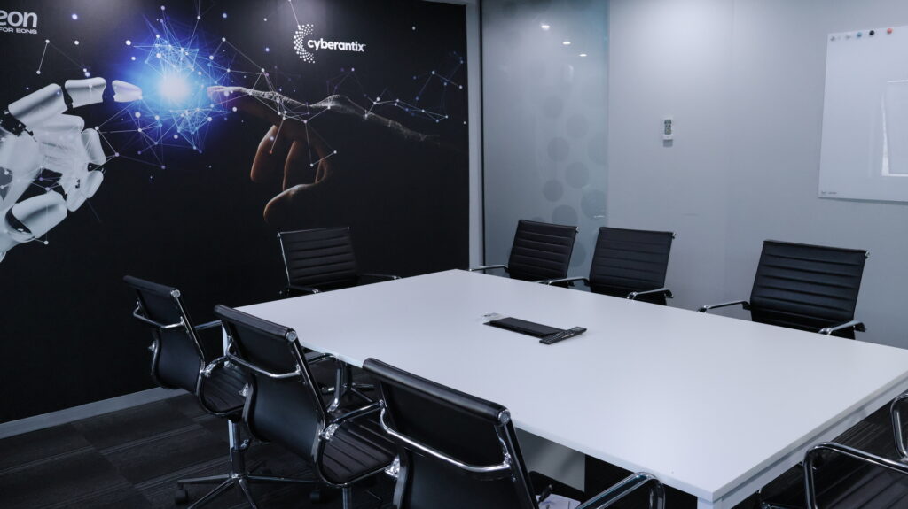 Modern conference room with a rectangular white table surrounded by six black leather office chairs with chrome frames. On the left wall, a large mural depicts a robotic hand reaching toward a human hand with a glowing network of connected nodes, alongside the text ‘cyberantix’ and ‘for eons.’ The opposite wall has a mounted whiteboard and a small control device, creating a sleek, professional workspace with a technology and innovation focus.