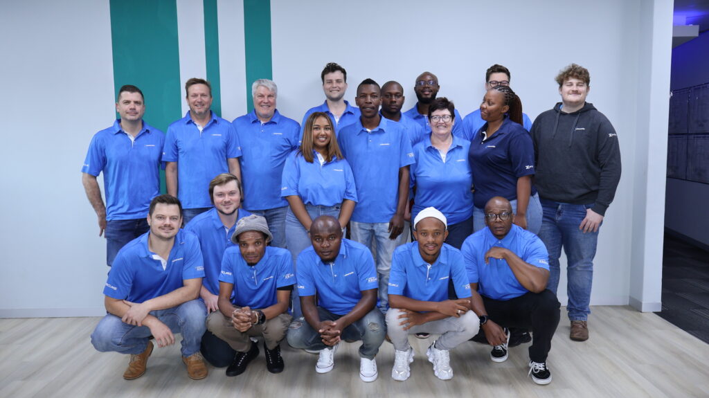 Group photo of sixteen individuals indoors, most wearing matching blue collared shirts with a small white logo, arranged in two rows with some standing and others kneeling or sitting in front. One person wears a dark blue shirt with a similar logo, and another wears a dark grey hoodie. The background features a light wall with two vertical green stripes and a light wood floor, conveying a professional team or staff group setting that highlights unity and collaboration found on the about us page.