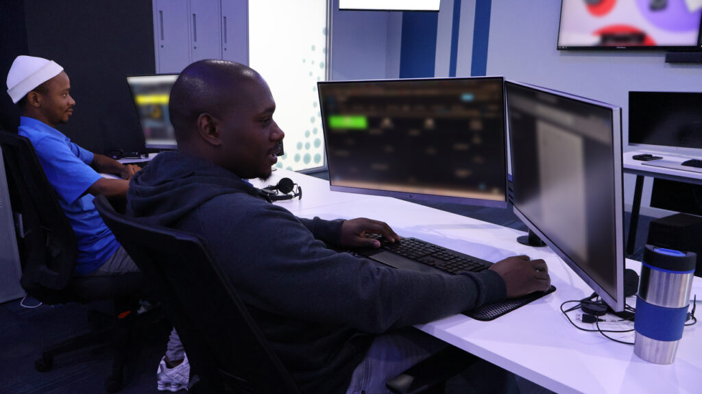 Two people seated at desks in a modern office, each working at dual‑monitor computer workstations. The person in the foreground wears a dark hoodie and uses a keyboard and mouse, while the person in the background in a blue shirt and white cap also works at a computer. The clean workspace features blue and white tones, blurred software interfaces on the monitors, and a stainless steel tumbler on the desk, representing a professional IT or programming environment found on the home page.